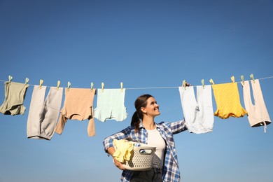 Photo of Smiling woman hanging baby clothes with clothespins on washing line for drying under blue sky outdoors