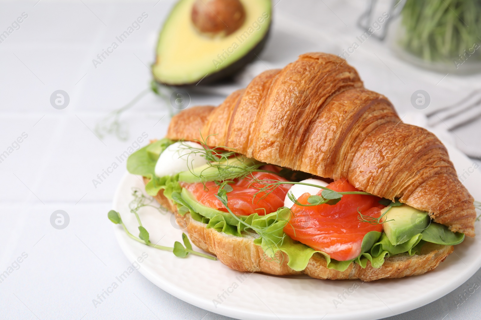 Photo of Tasty croissant with salmon, avocado, mozzarella and lettuce on white tiled table, closeup