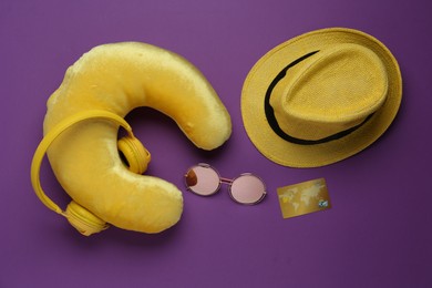 Photo of Yellow travel pillow with headphones, hat, sunglasses and credit card on purple background, flat lay