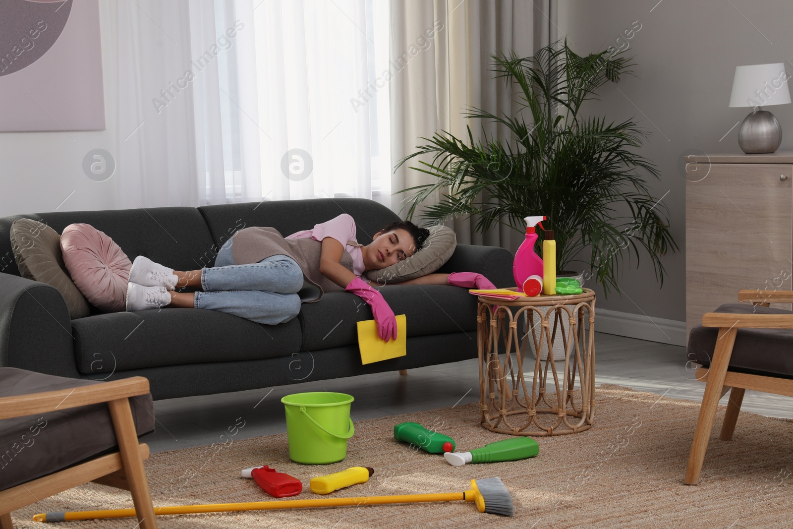 Photo of Tired young woman sleeping on sofa and cleaning supplies in living room