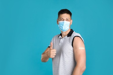 Photo of Vaccinated man with protective mask and medical plaster on his arm showing thumb up against light blue background. Space for text