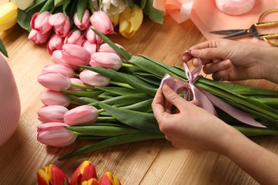 Photo of Woman making beautiful bouquet of fresh tulips and ribbon at wooden table, closeup