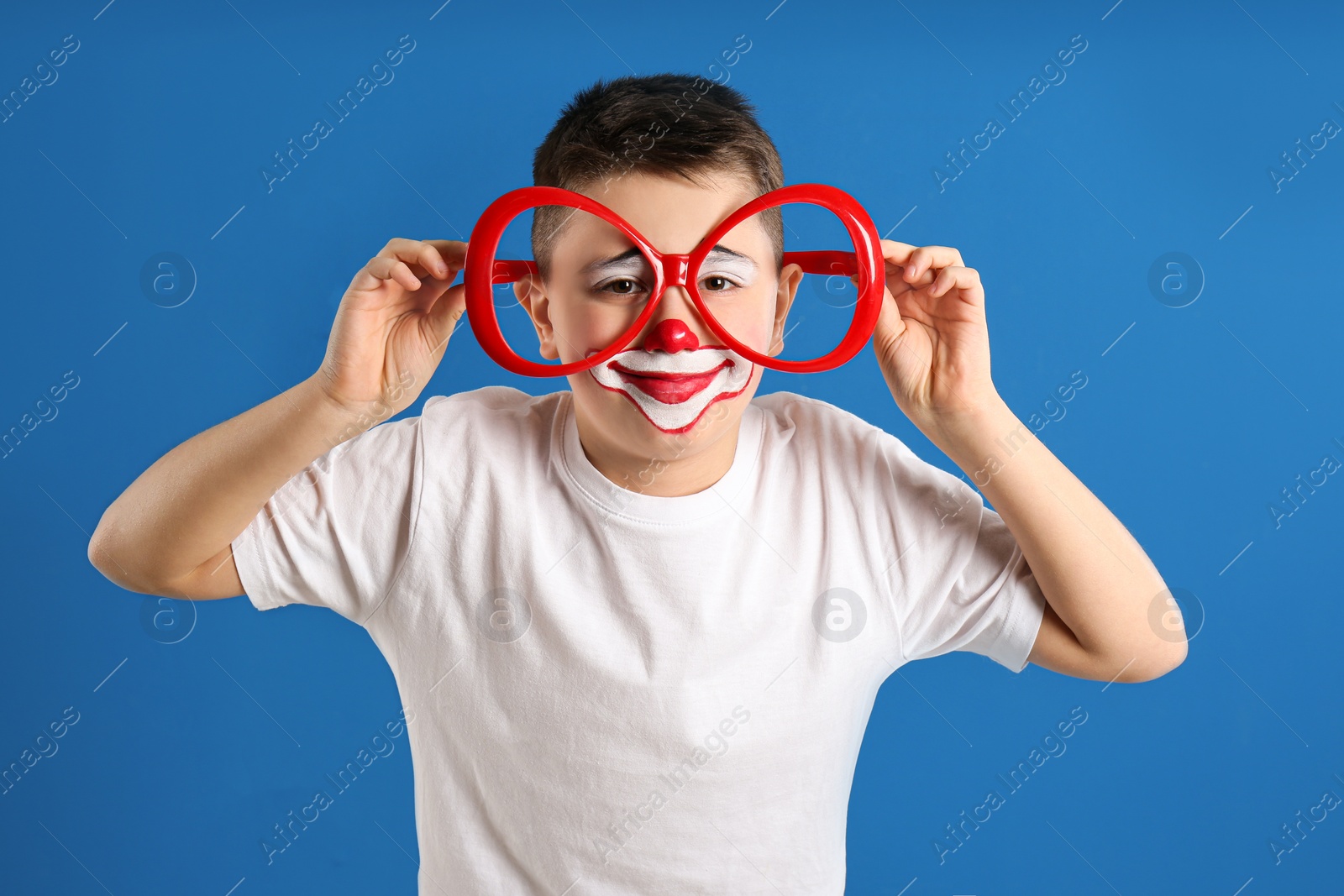 Photo of Preteen boy with clown makeup and party glasses on blue background. April fool's day