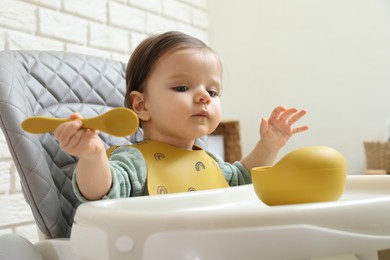 Photo of Cute little baby with spoon and bowl sitting in high chair indoors