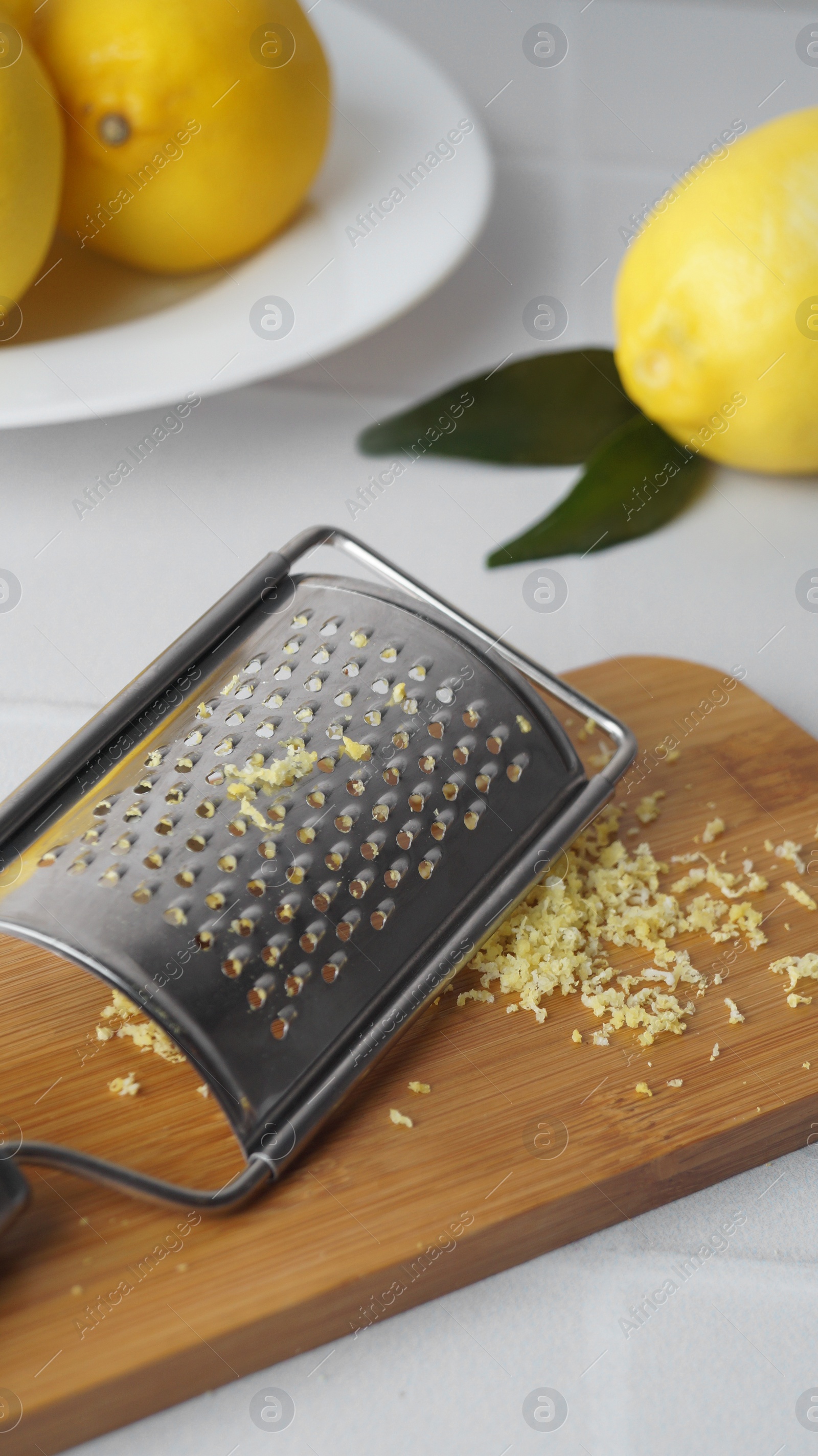 Photo of Wooden board with grater and fresh lemon zest on white tiled table, closeup