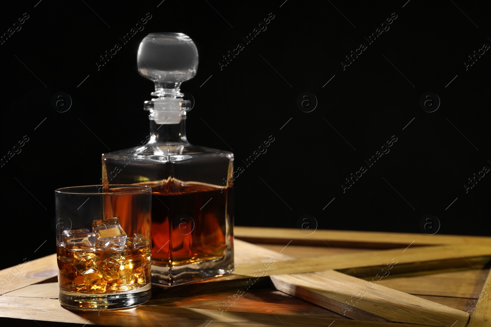 Photo of Whiskey with ice cubes in glass and bottle on wooden crate against black background, space for text