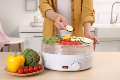 Woman putting cut zucchini into fruit dehydrator machine at wooden table in kitchen, closeup