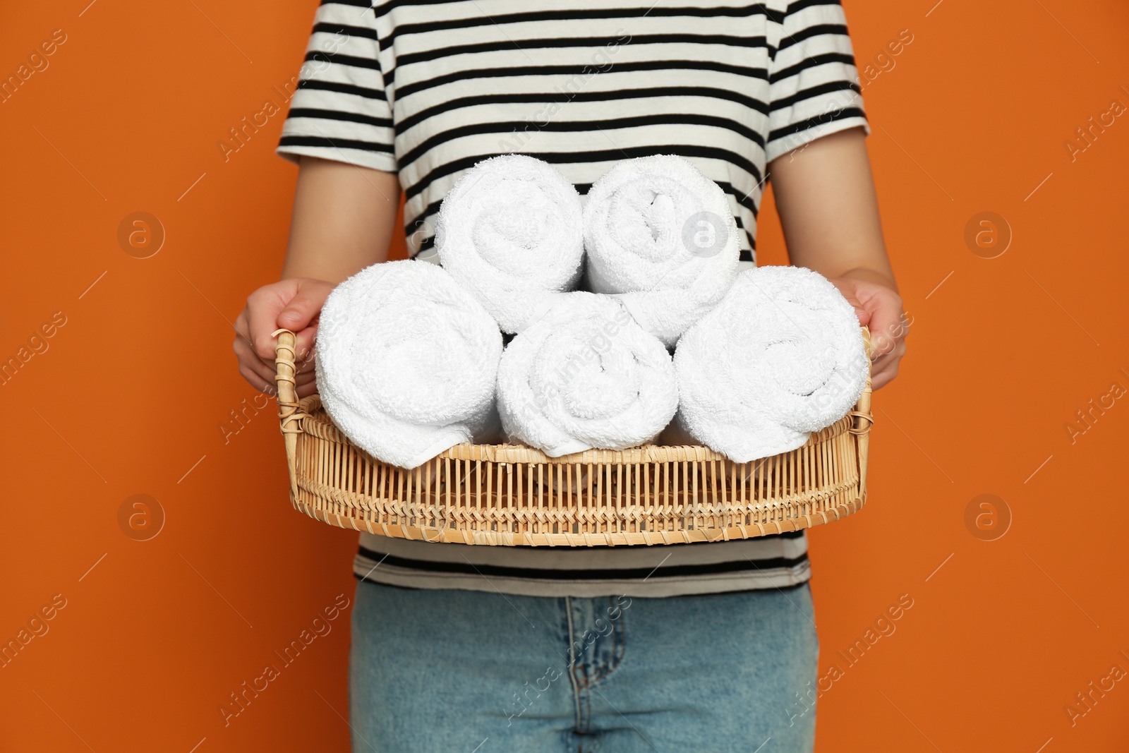 Photo of Woman holding wicker tray with rolled soft terry towels on orange background, closeup