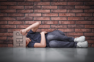 Photo of Little boy with sign HELP lying on floor near brick wall. Child in danger