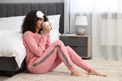 Beautiful young woman in stylish pyjama and sleep mask with cup of drink on floor at home