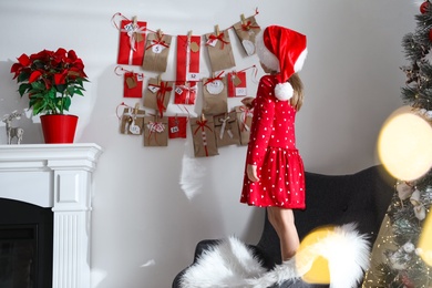 Photo of Cute little girl in Santa hat taking gift from Christmas advent calendar at home