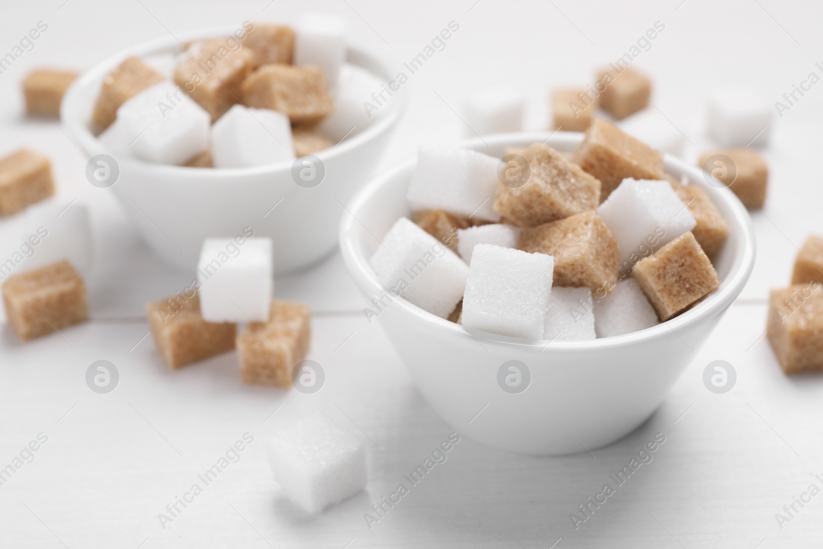 Photo of Different sugar cubes in bowls on white table, closeup