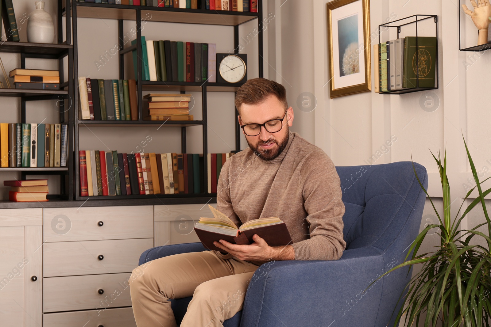 Photo of Young man reading book in armchair indoors. Home library