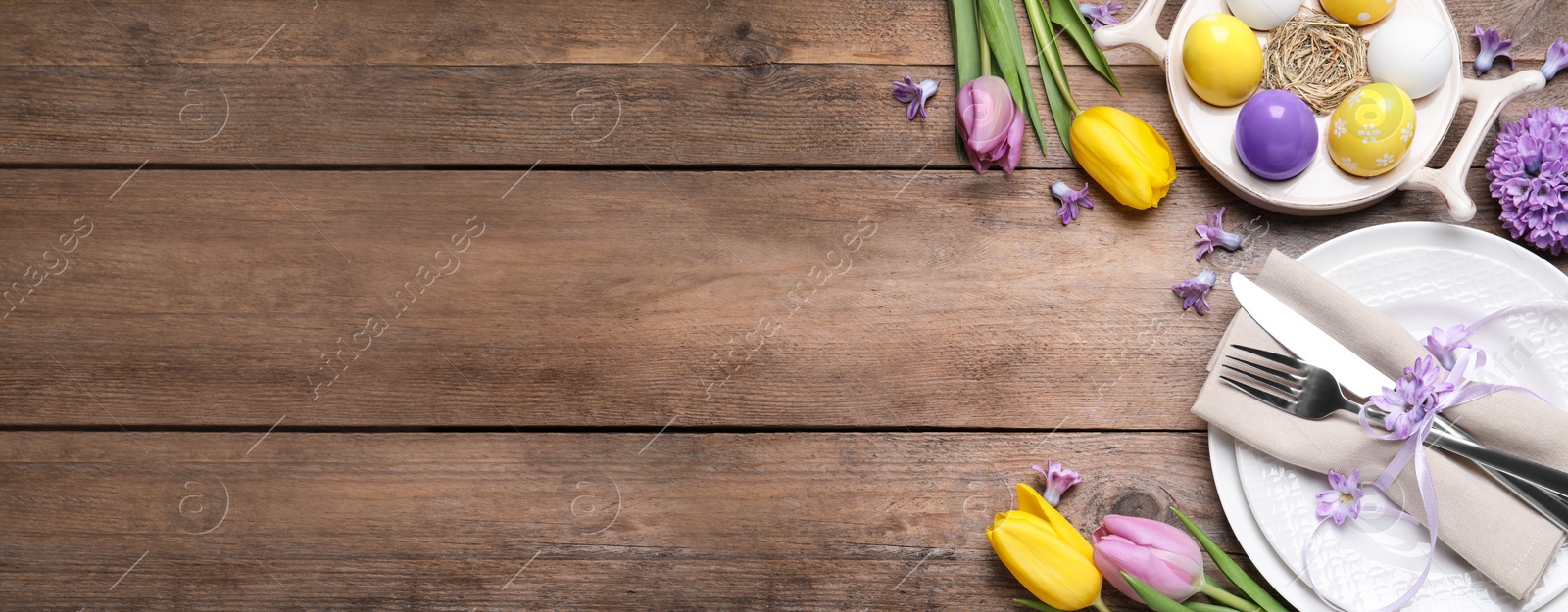 Image of Festive Easter table setting with painted eggs and flowers on wooden background, flat lay. Space for text