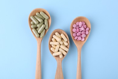 Different vitamin pills in wooden spoons on light blue background, flat lay