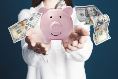 Image of Young woman holding piggy bank and American dollars falling on color background, closeup 