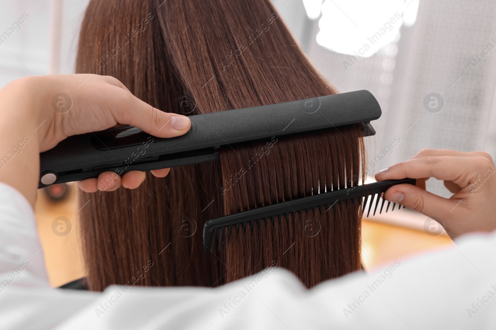 Photo of Hairdresser straightening woman's hair with flat iron in salon, closeup