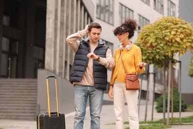 Photo of Being late. Worried man and woman with suitcase looking at watch outdoors