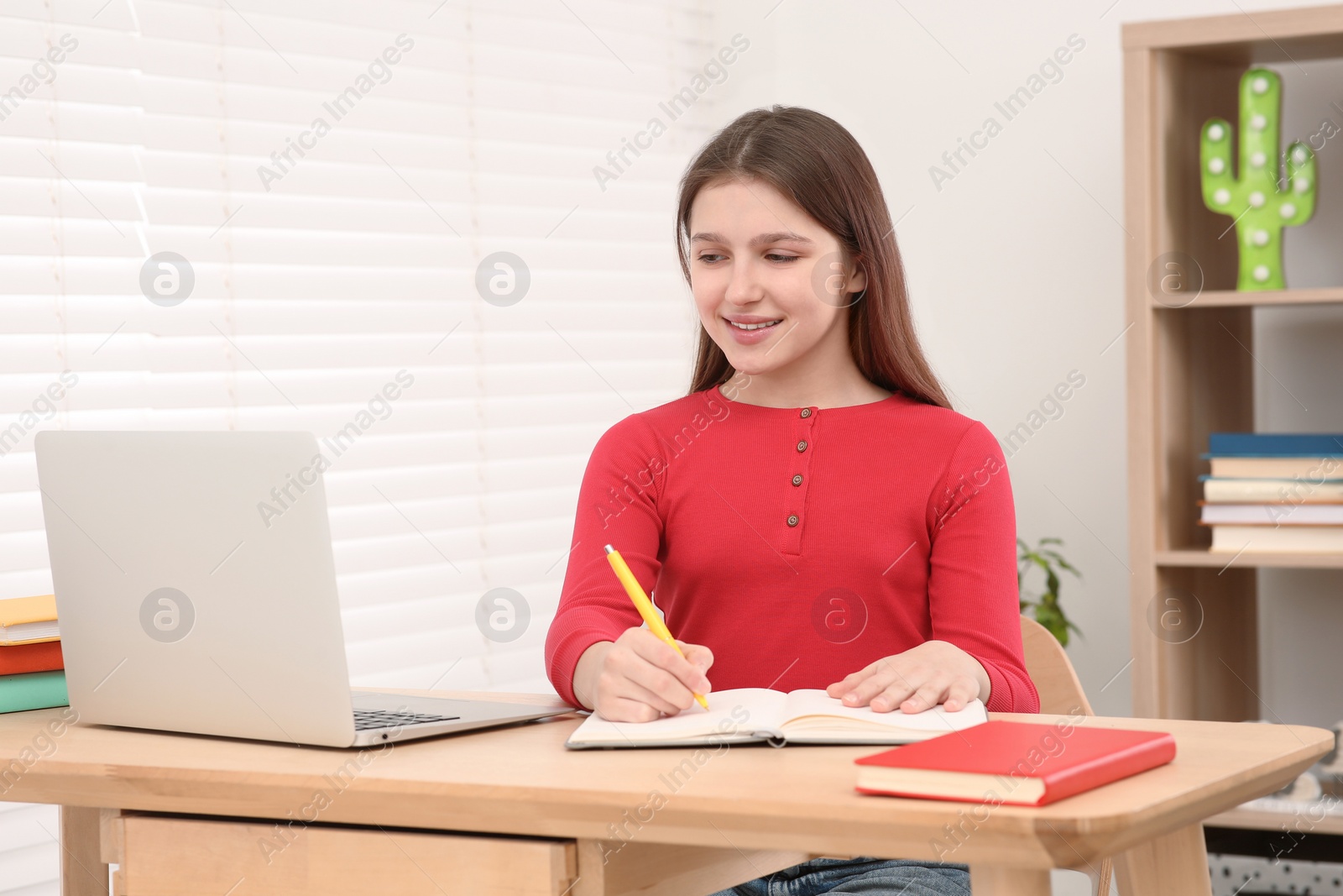 Photo of Cute girl writing in notepad near laptop at desk in room. Home workplace