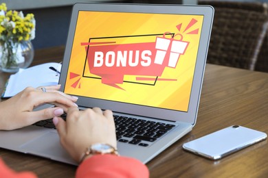 Bonus gaining. Woman using laptop at wooden table indoors, closeup. Illustration of gift box and word on device screen