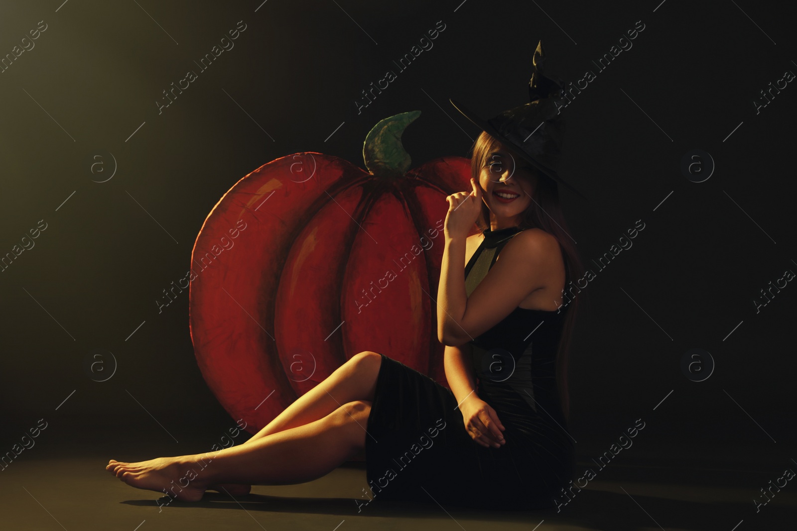 Photo of Young woman wearing witch costume near decorative pumpkin on black background. Halloween party