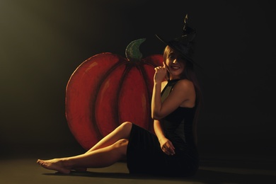 Young woman wearing witch costume near decorative pumpkin on black background. Halloween party