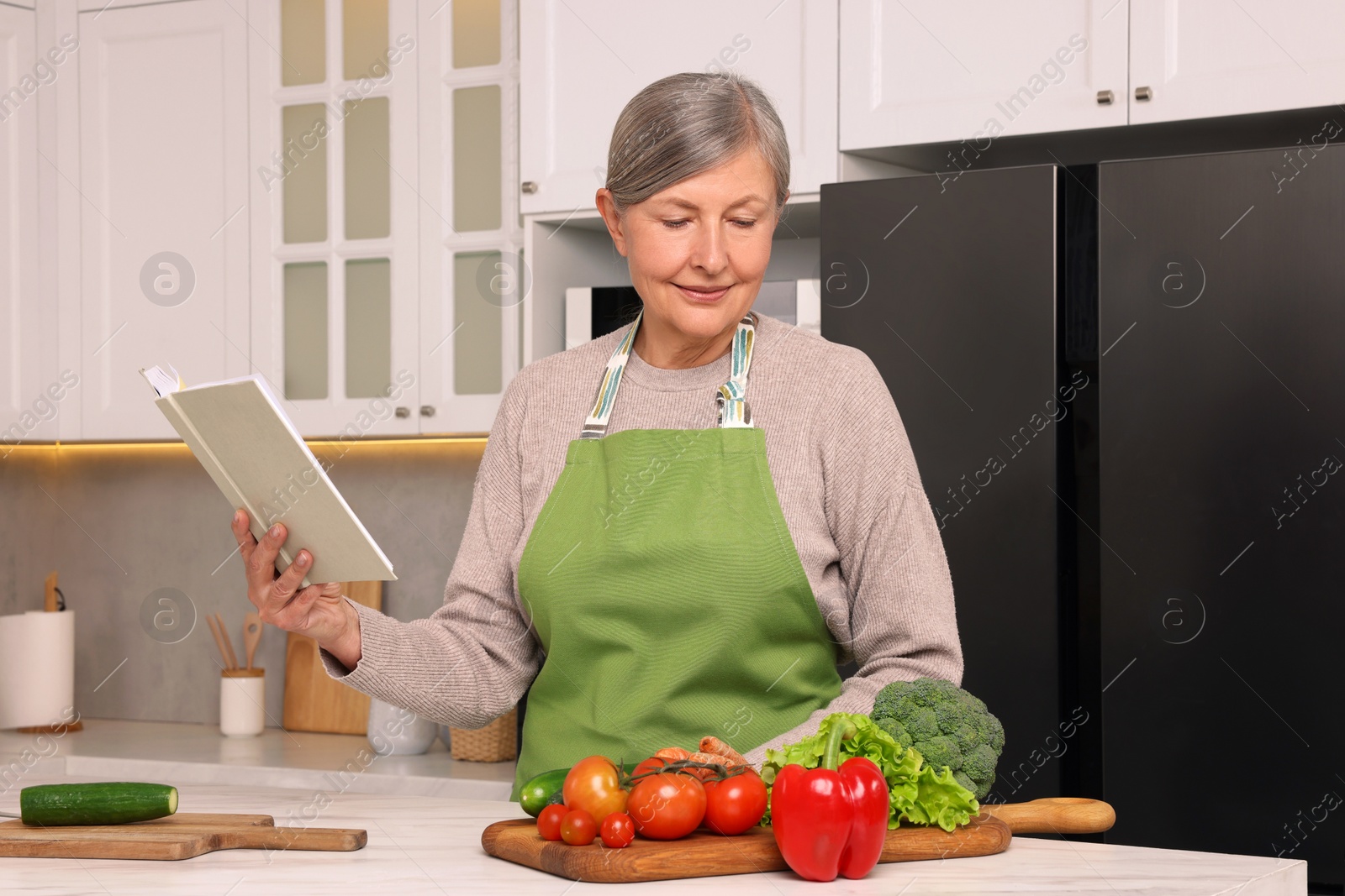 Photo of Senior woman with recipe book and products at table in kitchen