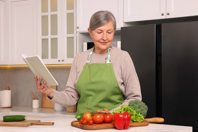 Photo of Senior woman with recipe book and products at table in kitchen