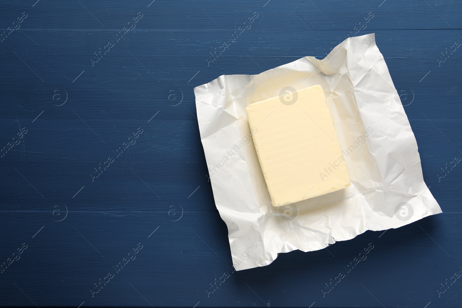 Photo of Block of tasty butter in open foil packaging on blue wooden table, top view. Space for text