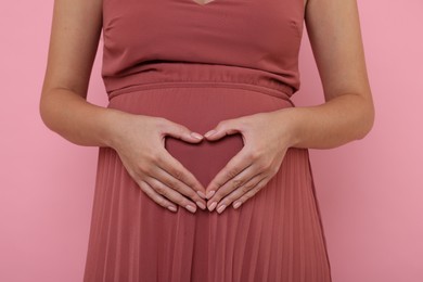 Photo of Pregnant woman in dress making heart with hands on pink background, closeup
