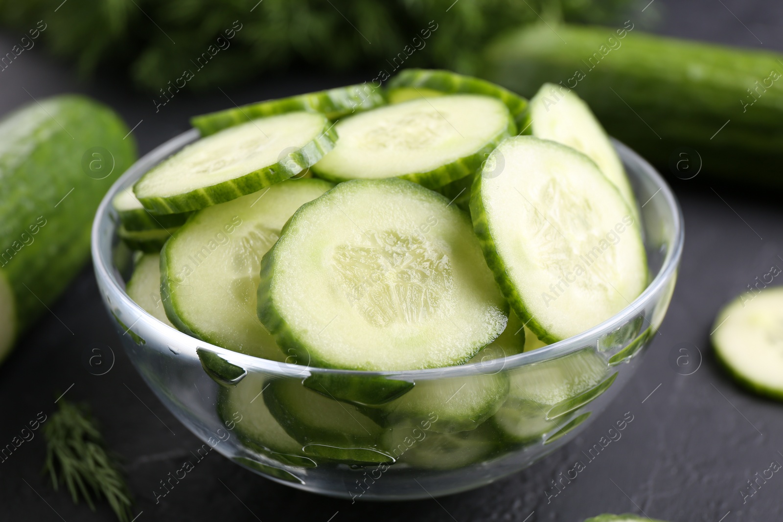 Photo of Cut cucumber in glass bowl on dark gray textured table, closeup