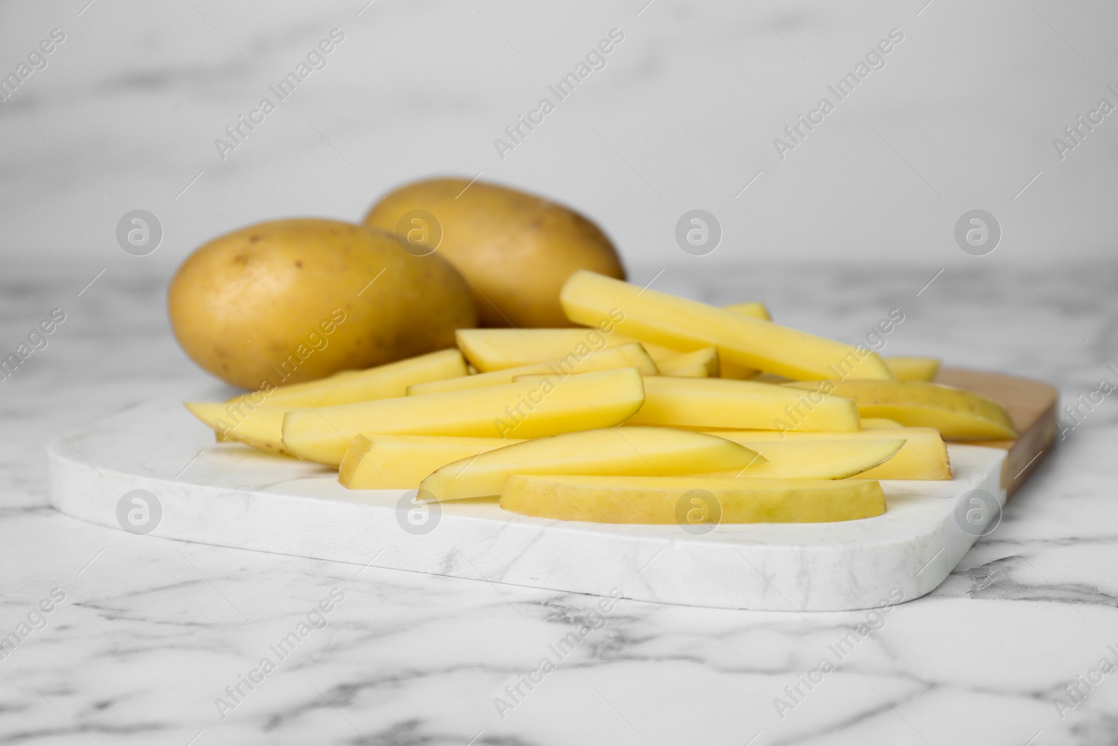 Photo of Whole and cut raw potatoes on white marble table, closeup. Cooking delicious French fries