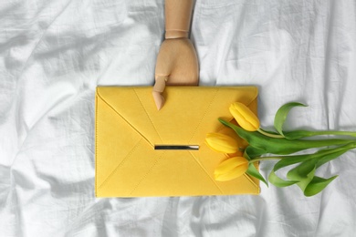 Photo of Stylish envelope bag with spring tulips and wooden mannequin hand on crumpled sheet, flat lay