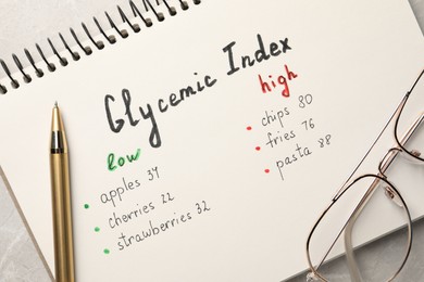 Photo of List with products of low and high glycemic index in notebook, pen and glasses on table, top view
