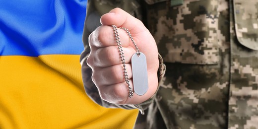 Closeup view of soldier in uniform holding military ID tag and Ukrainian flag on background. Banner design