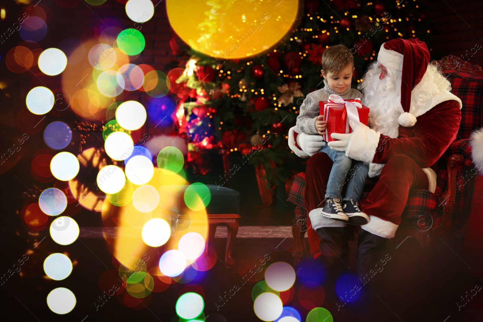 Photo of Santa Claus and little boy with gift near Christmas tree indoors