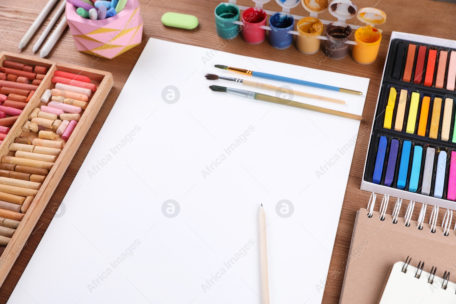 Photo of Blank sheet of paper, colorful chalk pastels and other drawing tools on wooden table. Modern artist's workplace