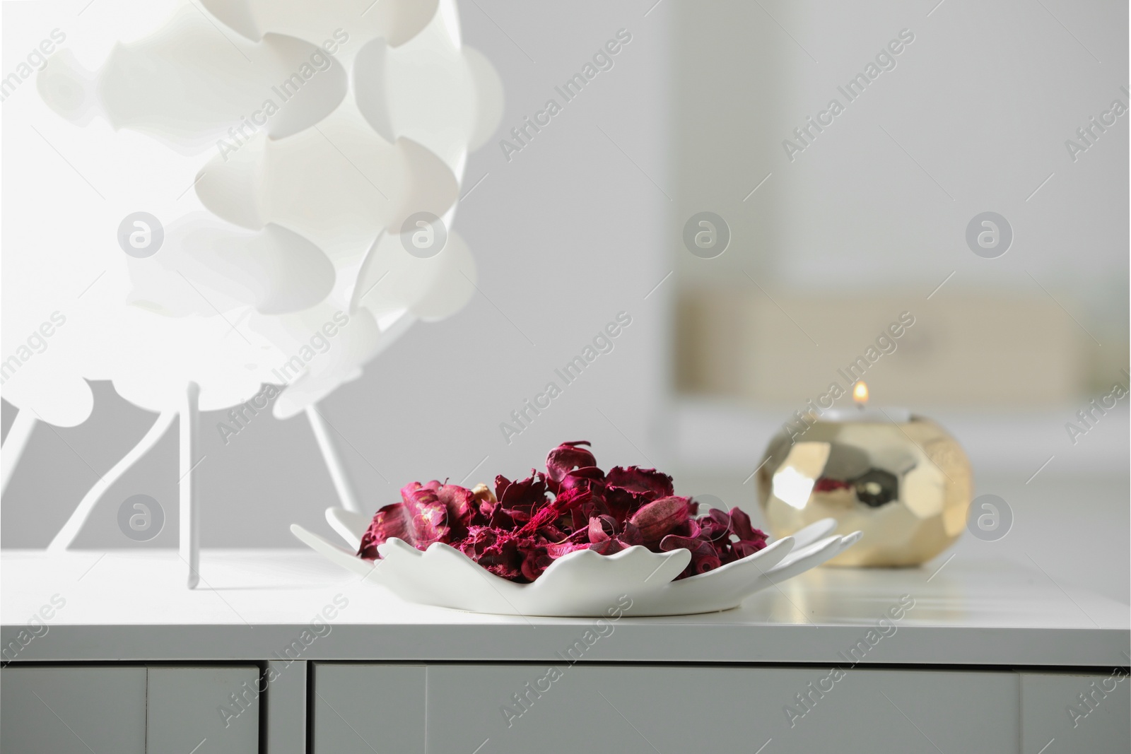Photo of Plate with aromatic potpourri, lamp and burning candle on white chest of drawers in room