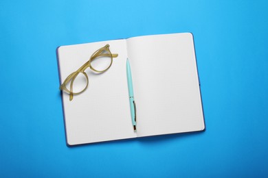 Open notebook, pen and glasses on light blue background, top view