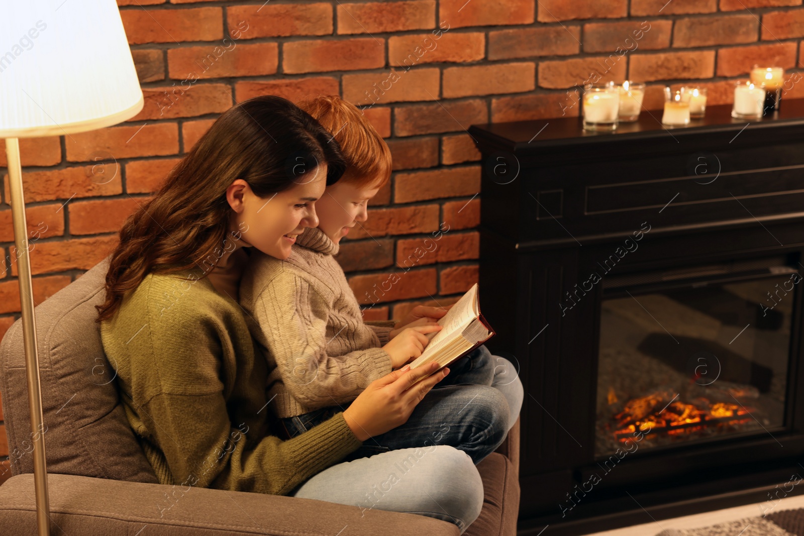 Photo of Happy mother and son reading book together near fireplace at home