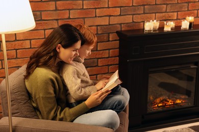 Photo of Happy mother and son reading book together near fireplace at home
