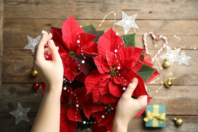 Photo of Woman decorating poinsettia (traditional Christmas flower) with elegant bead garland at wooden table, top view