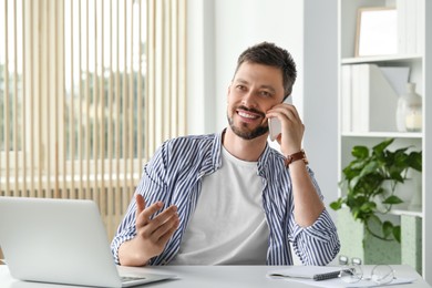 Photo of Happy man talking on smartphone while working with laptop in office