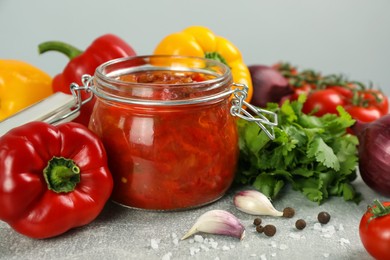 Photo of Glass jar of delicious canned lecho and fresh ingredients on light grey table, closeup