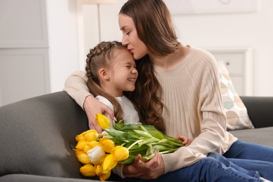 Photo of Happy woman kissing her cute daughter with bouquet of tulips on sofa at home