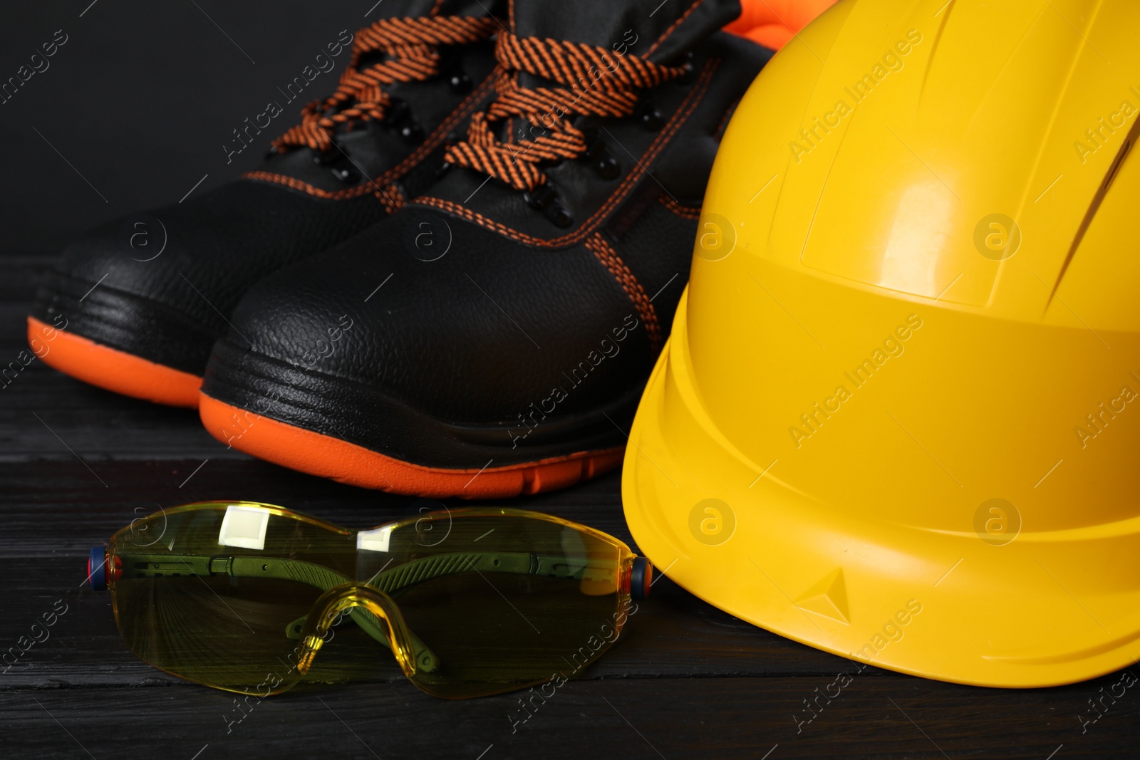 Photo of Pair of working boots, hard hat and goggles on black wooden surface, closeup