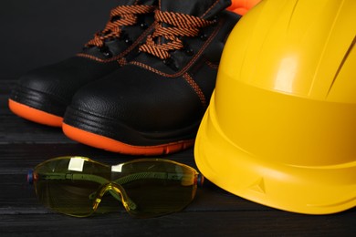 Photo of Pair of working boots, hard hat and goggles on black wooden surface, closeup
