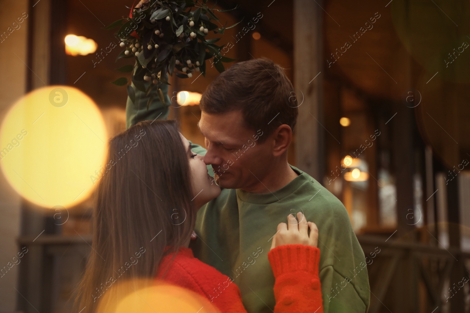 Photo of Happy couple kissing under mistletoe bunch outdoors, bokeh effect