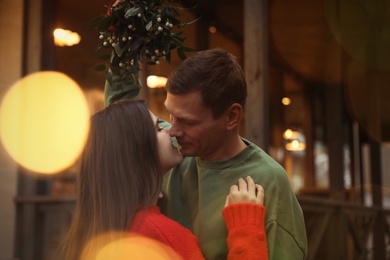 Photo of Happy couple kissing under mistletoe bunch outdoors, bokeh effect