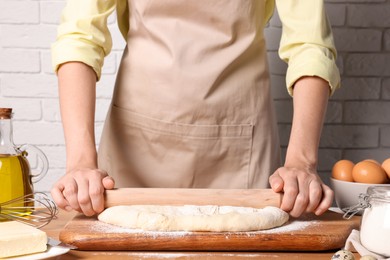 Photo of Woman rolling dough with wooden pin at table near white brick wall, closeup
